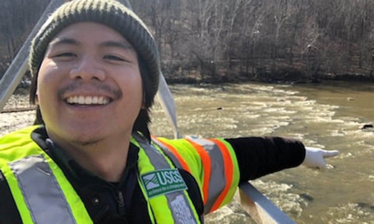 Adult man wearing a neon green and orange safety vest standing in front of a river.