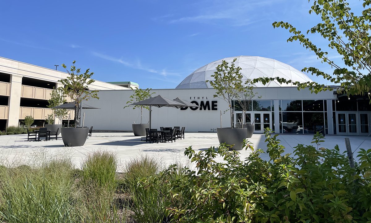 An image of the Science Museum's Dome Plaza showing black recycled plastic tables with a dark umbrellas shading them. The words "The Dome" are visible on the wall in the background. The concrete is bright white.