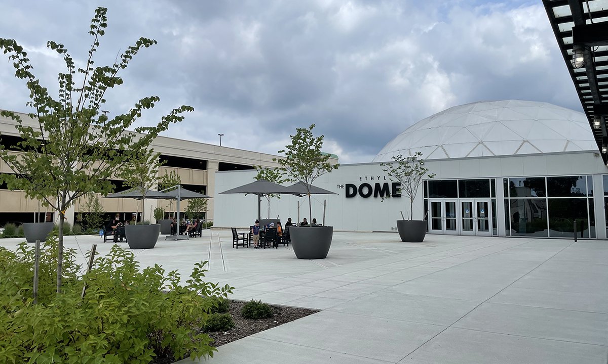 An image of the Science Museum's Dome Plaza showing black recycled plastic tables with a dark umbrellas shading them. The words "The Dome" are visible on the wall in the background. The concrete is bright white.