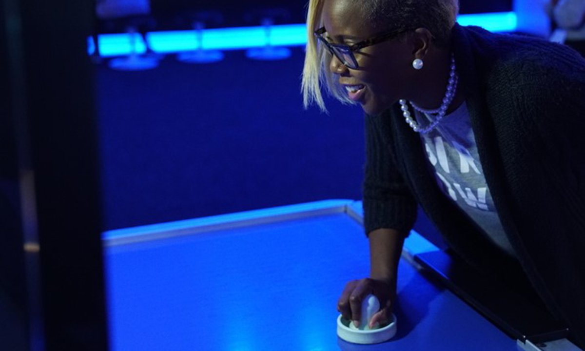 A guest playing air hockey against a robot. She is leaned over the table with one hand on the plastic disk, having just hit the puck.