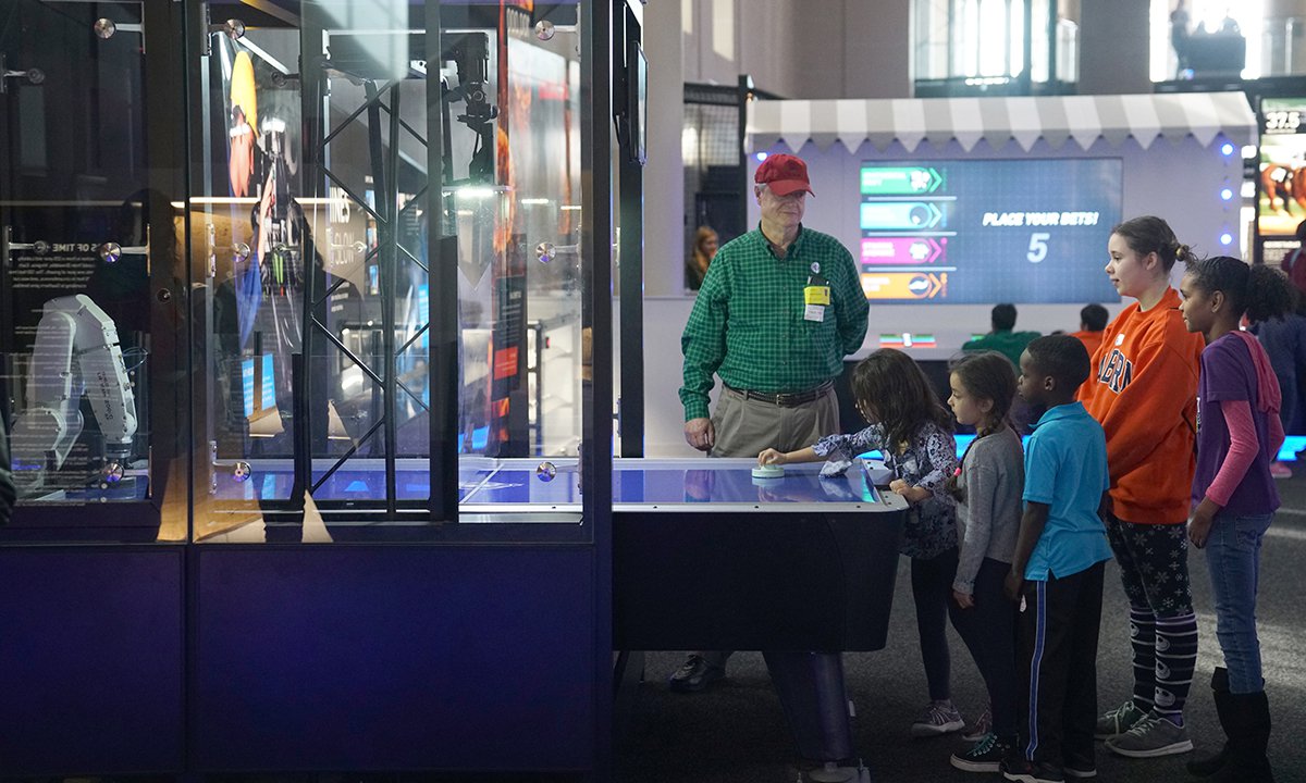 A photo of the air hockey robot table. A guest plays while children and one adult stand around watching.