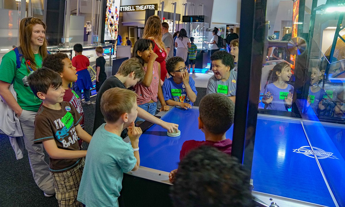 A school group play air hockey against the robot. One child plays while about a dozen other children and one adult stand around the table watching.